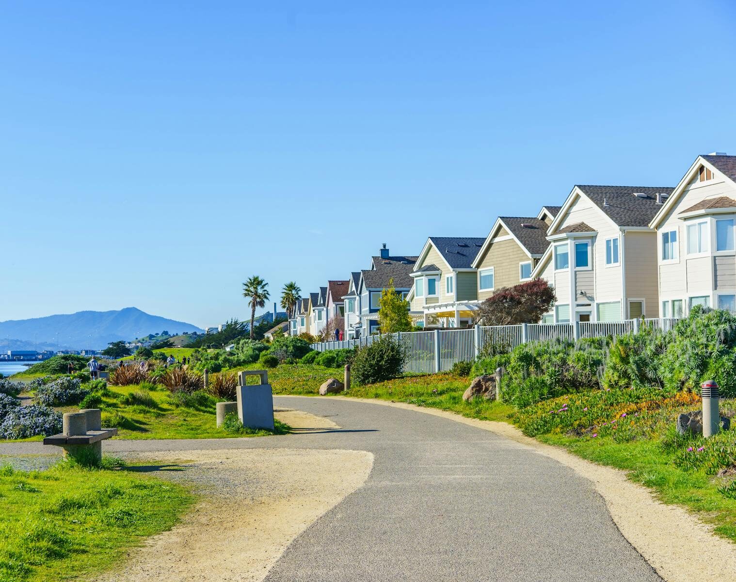 Bright daytime view of a coastal neighborhood with a scenic path and distant mountain backdrop.