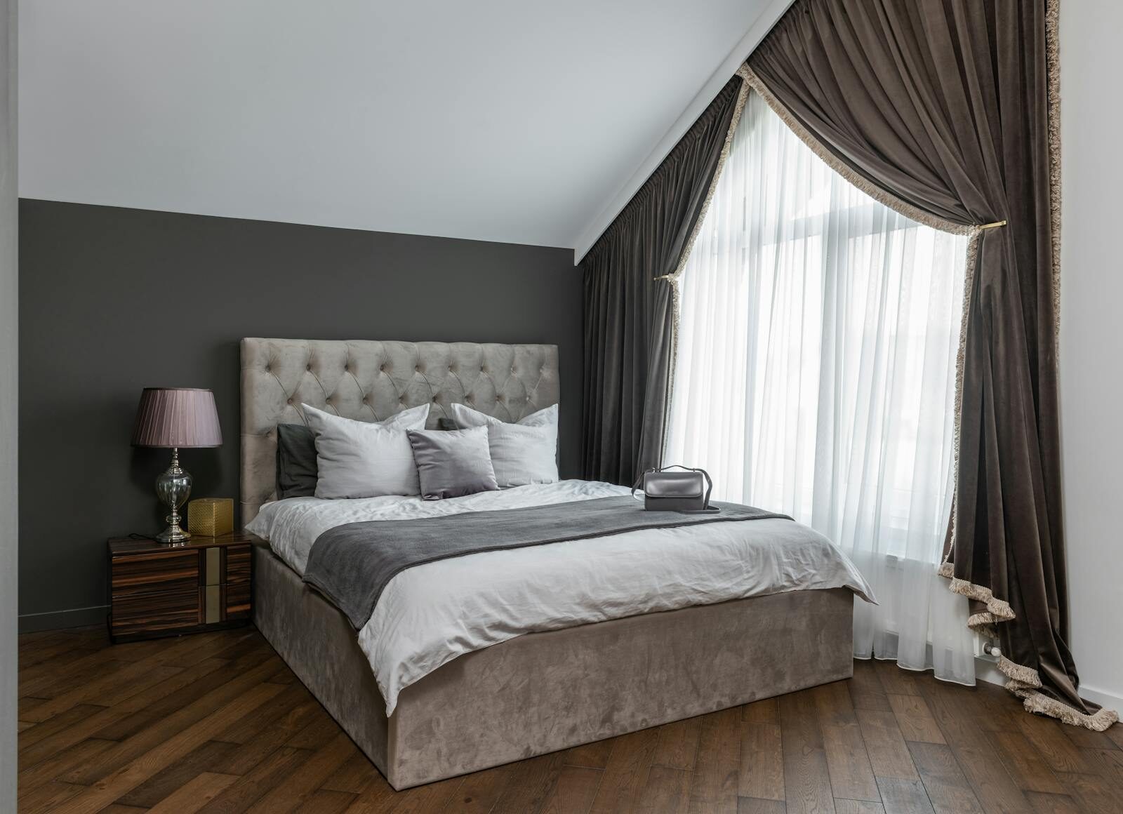 Comfortable bed with blanket and cushions placed on parquet near window with curtains and lamp on bedside table in modern bedroom