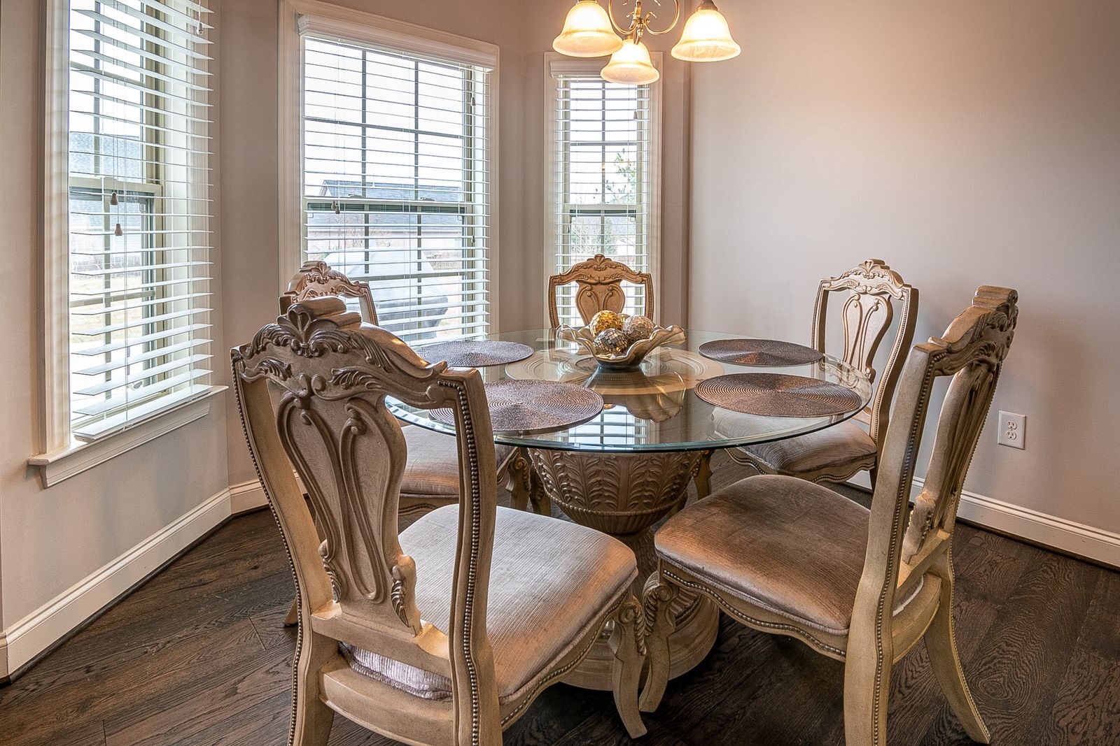 A well-lit dining room featuring classic wooden chairs and a glass table set against tall windows.