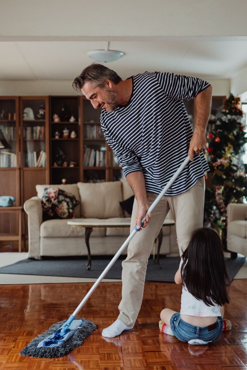 A smiling father mops the floor with his daughter nearby, creating joyful memories.