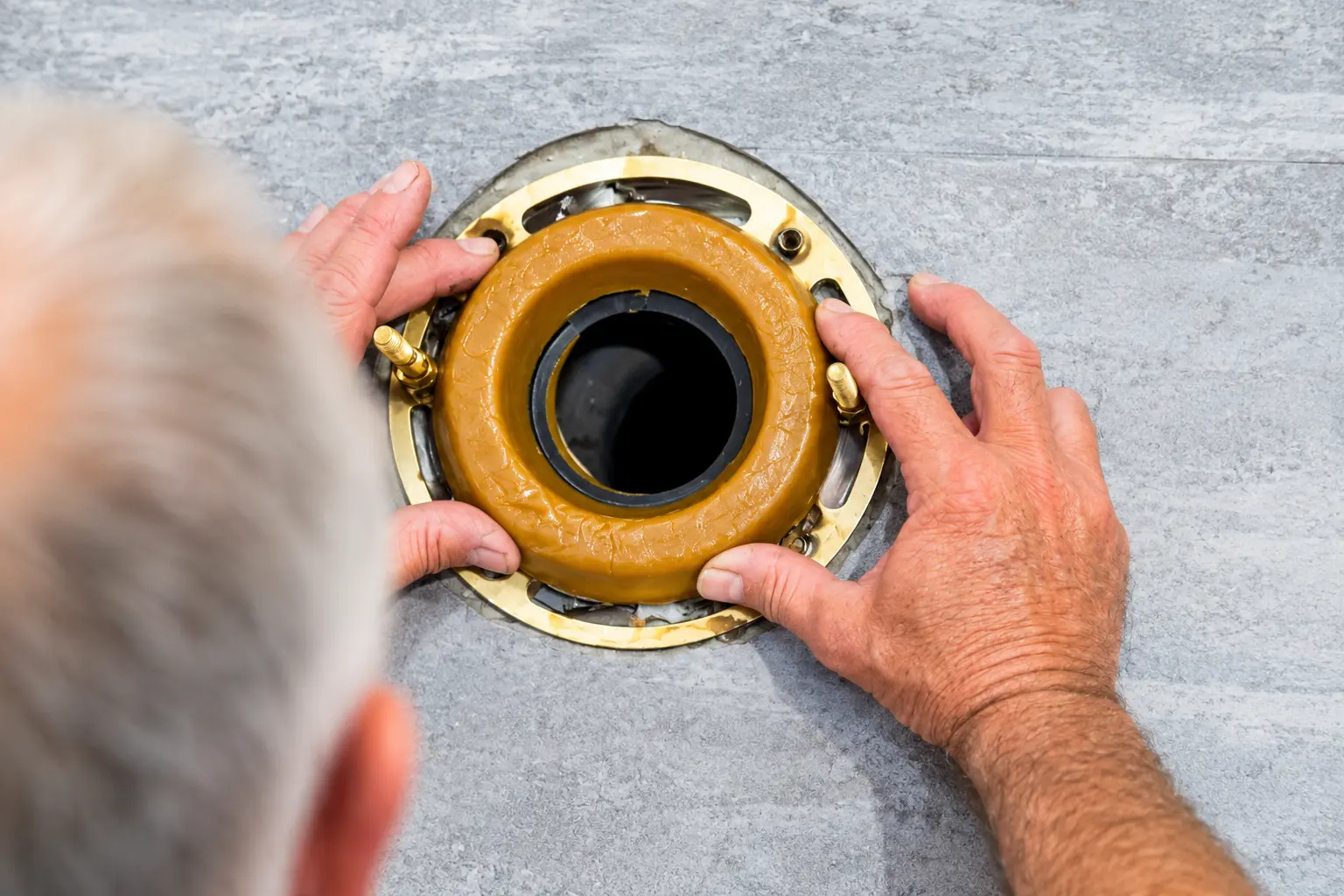 Person installing a toilet wax ring gasket over a brass toilet flange and drain opening on a gray floor