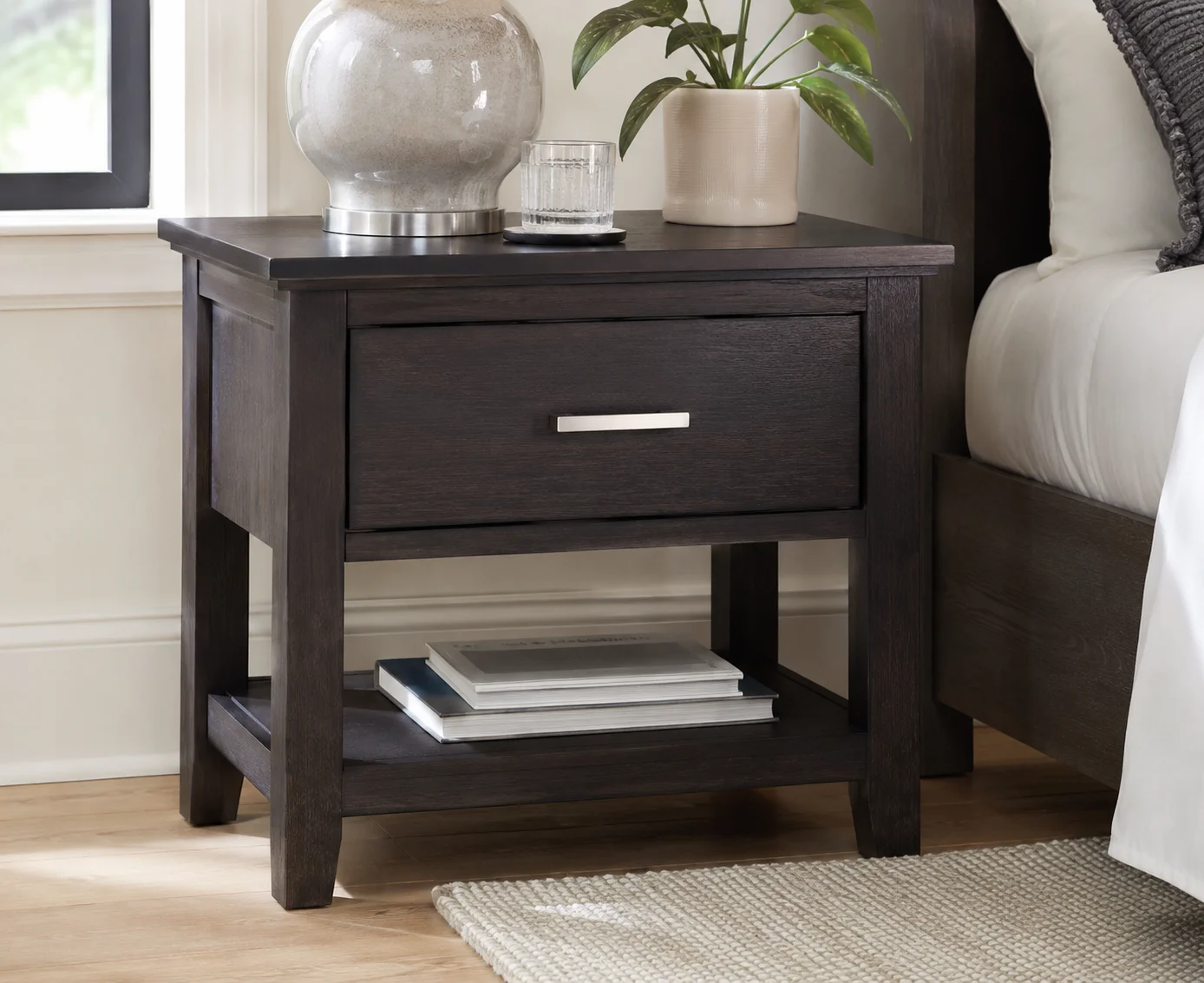 Ashley Finch One-Drawer Nightstand in dark brown wood beside a modern bed, styled with a lamp, plant, and books.