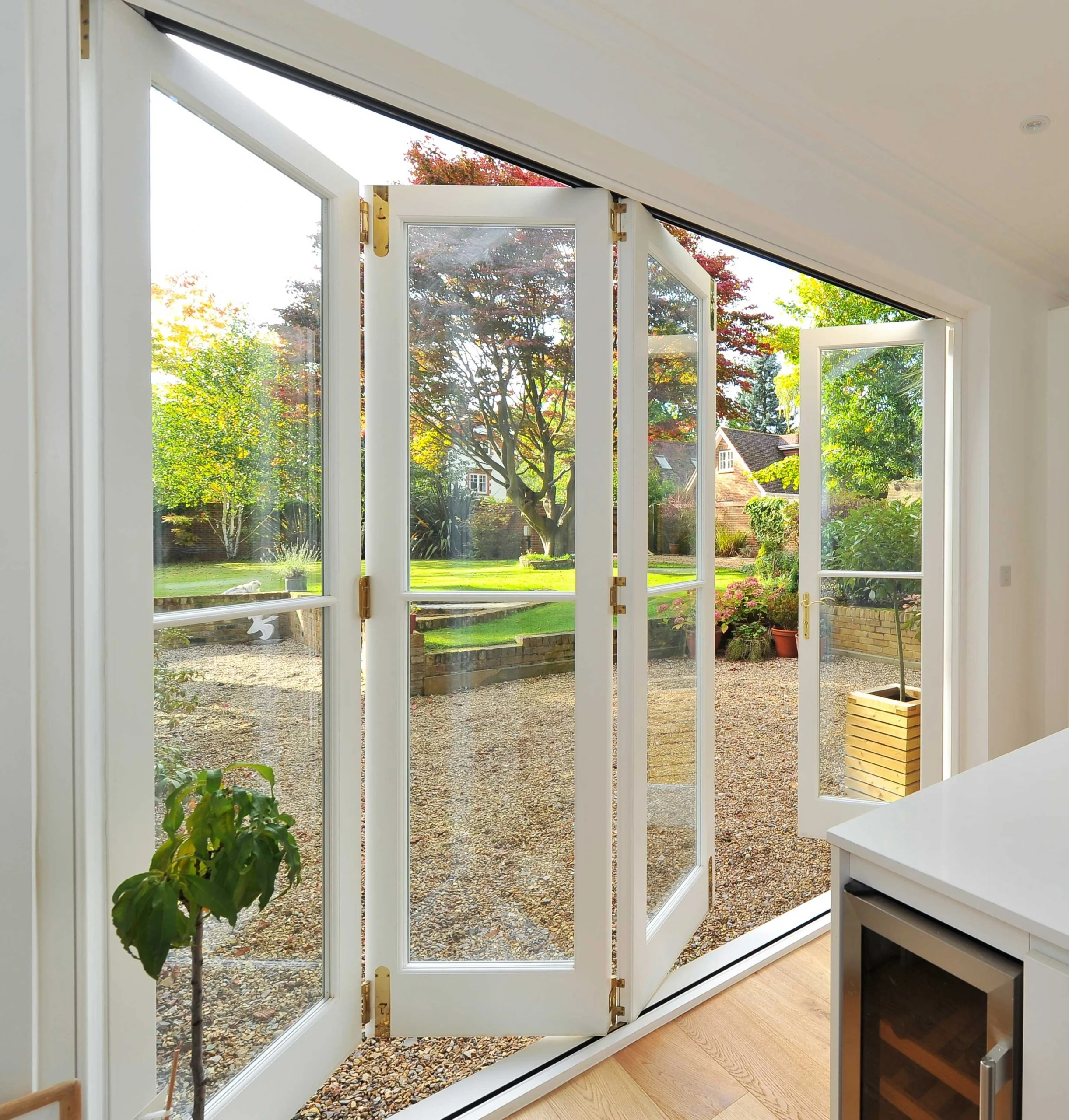 White bifold glass doors folded open to a sunny garden with green lawn, colorful flowers, gravel patio, and warm hardwood flooring in a modern home.